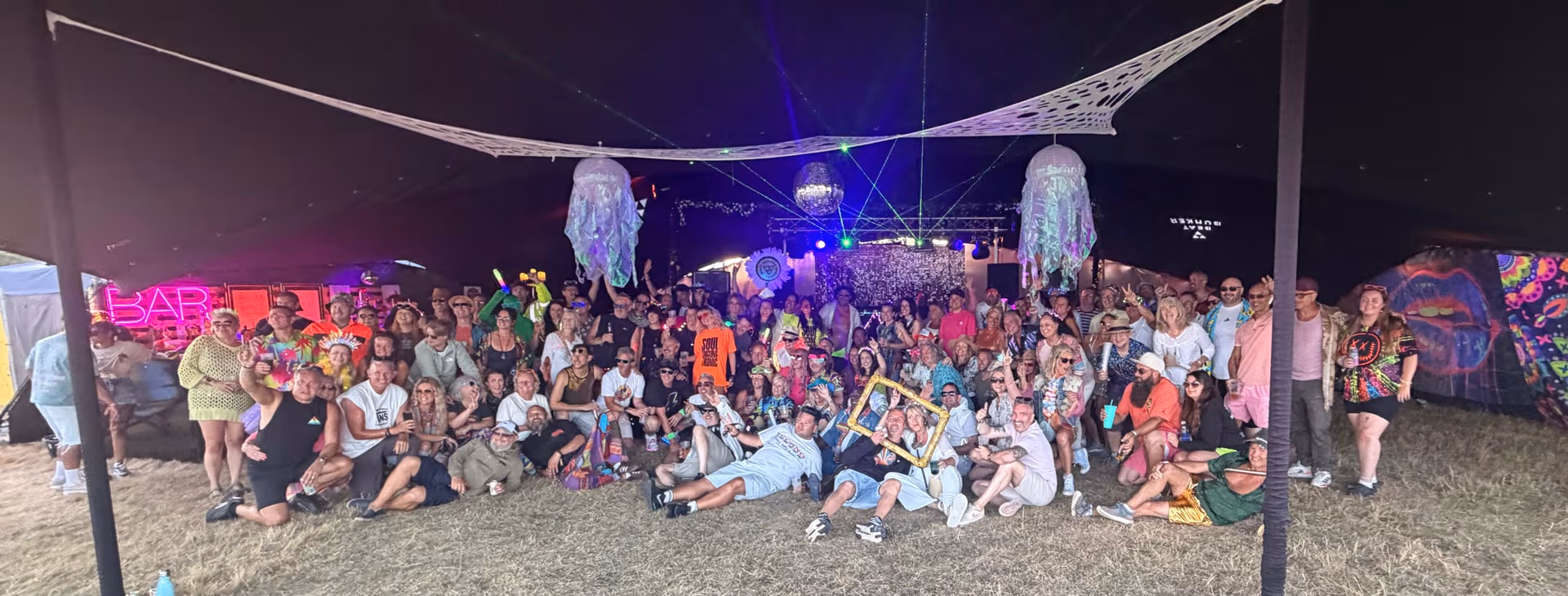 The Beat Bunker 2026 crowd posing together under the stretch tent, lit by neon, lasers and disco balls.